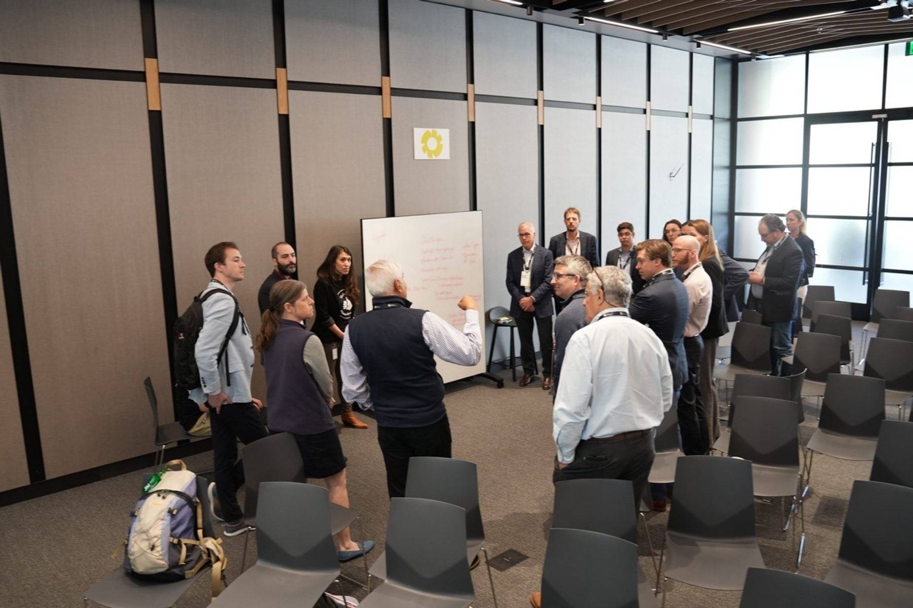 People stand in a circle in The Engine's event space to hear a man in a blue vest speak.