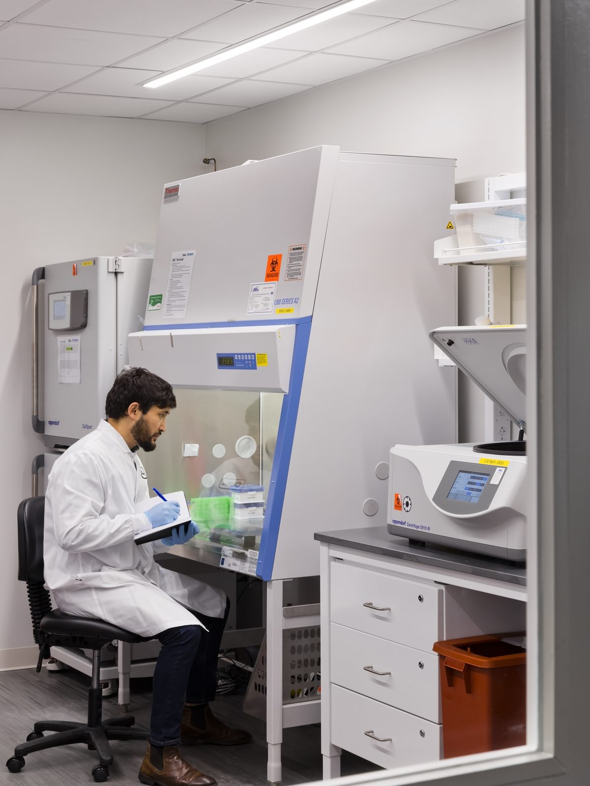 A scientist in a lab coat takes notes on an experiment in a fume hood in The Engine's BSL-2 wet lab space.